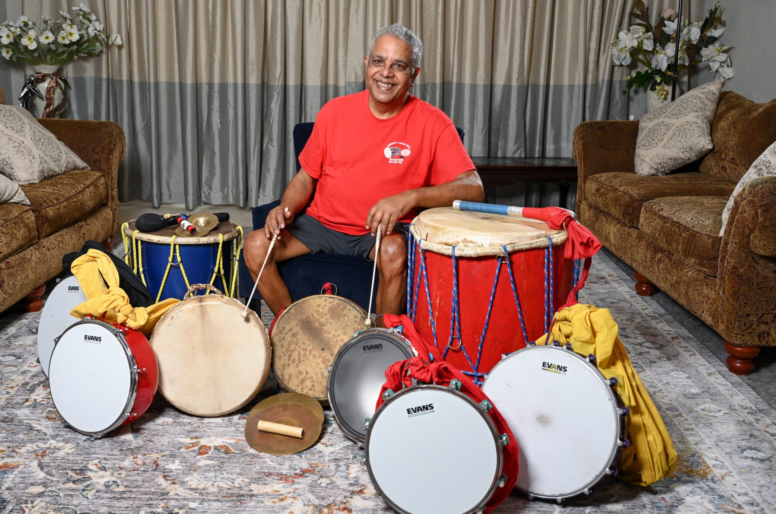 Romeo Ragbir sits surrounded by drums of various sizes. He holds a drum stick in each hand.