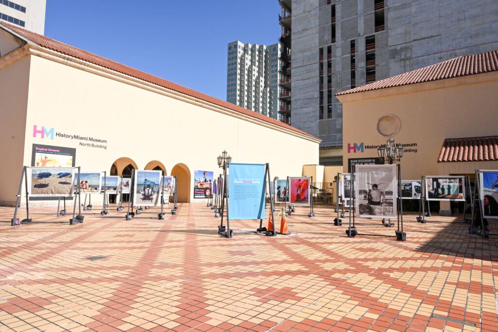 View of Museum of Miami from the Cultural Plaza. Two beige buildings sit on an orange and yellow tiled plaza. A photo exhibit displayed on wooden frames sit between them. In the distance, a building is being constructed against a blue sky.