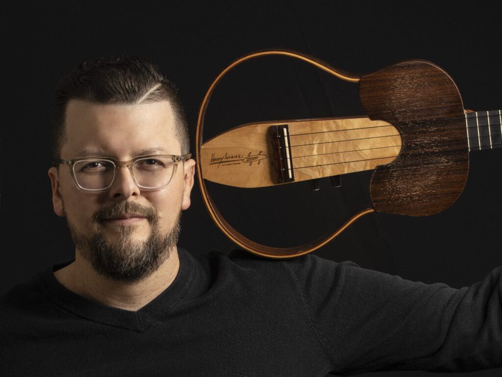Henry Linarez wears a black shirt and holds his cuatro guitar over his shoulder while looking into the camera