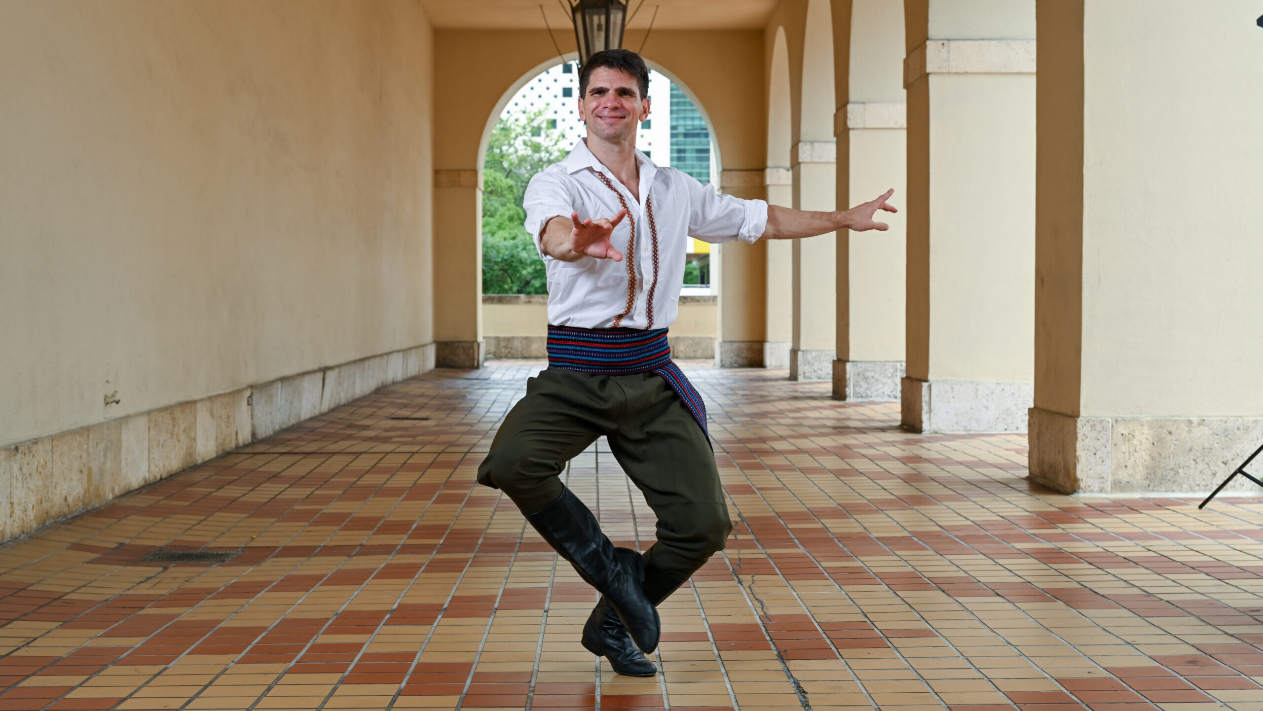 A dancer poses with his toes pointed towards the center and his arms outstretched. He outdoors in a corridor with hanging lamps and is wearing a white shirt with embroidered designs, green pants, and a colorful belt