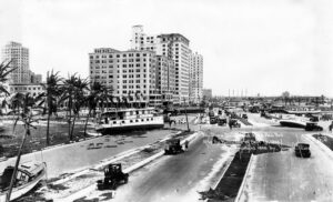 A black and white image of a street view in downtown Miami in the aftermath of a hurricane. Boats can be seen strewn along the street next to the bay with downed poles nearby.