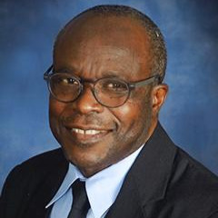 A person wearing round glasses and a suit smiles against a blue backdrop.