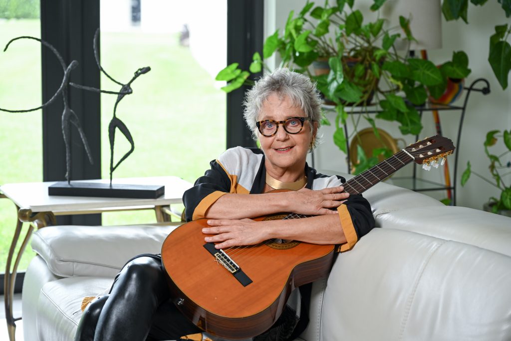 A woman (Alejandra Czarny) with short grey hair and glasses sits on a couch holding a guitar and smiling at the camera