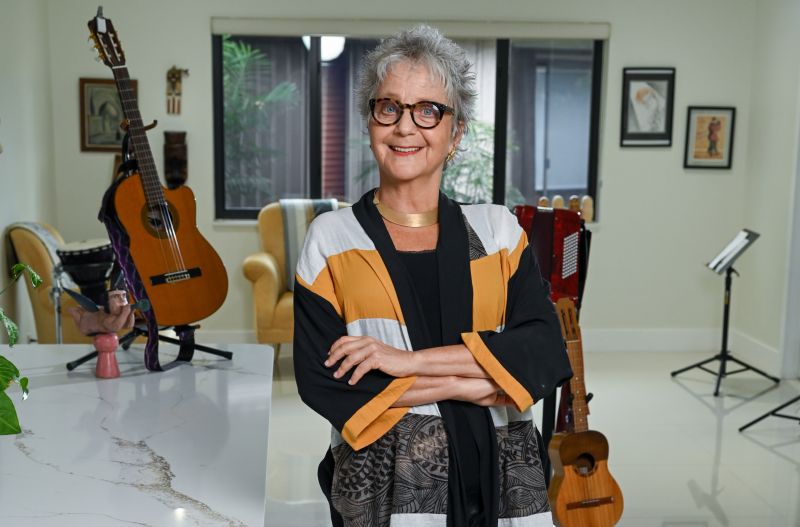 A woman (Alejandra Czarny) stands with her arms crossed and smiling at the camera. Behind her are two guitars and a music stand