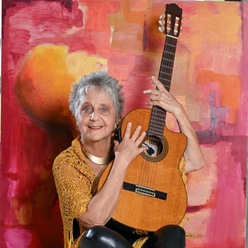 A woman (Alejandra Czarny) smiles at the camera while holding a guitar in front of a red and orange backdrop