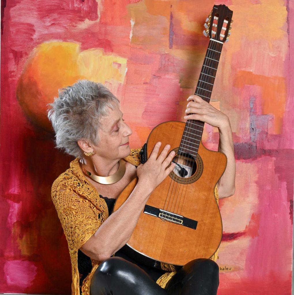 A woman (Alejandra Czarny) sits on the floor with her legs crossed, holding a guitar in front of her. She is holding the body and neck of the guitar as she smiles and looks at it