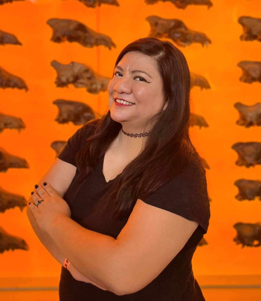 A person stands with their arms crossed in front of a large museum exhibition case with several rows of brown dire wolf skulls against an orange backdrop.