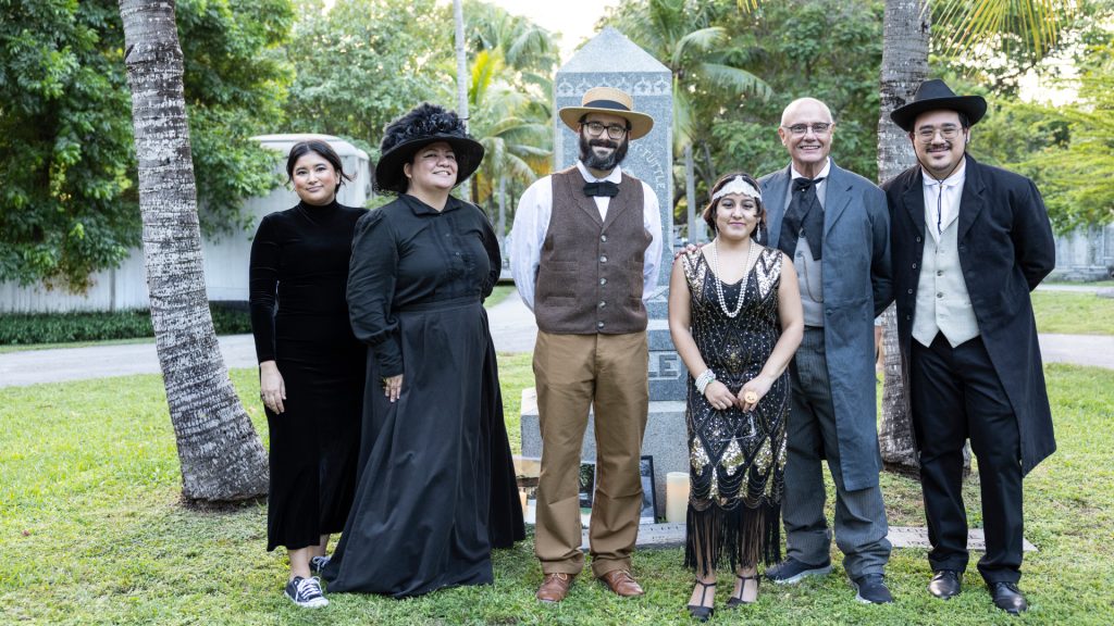 A group of people dressed in historical costumes stand together in front of a tall headstone inside of a cemetery. Green grass and trees can be seen in the background.