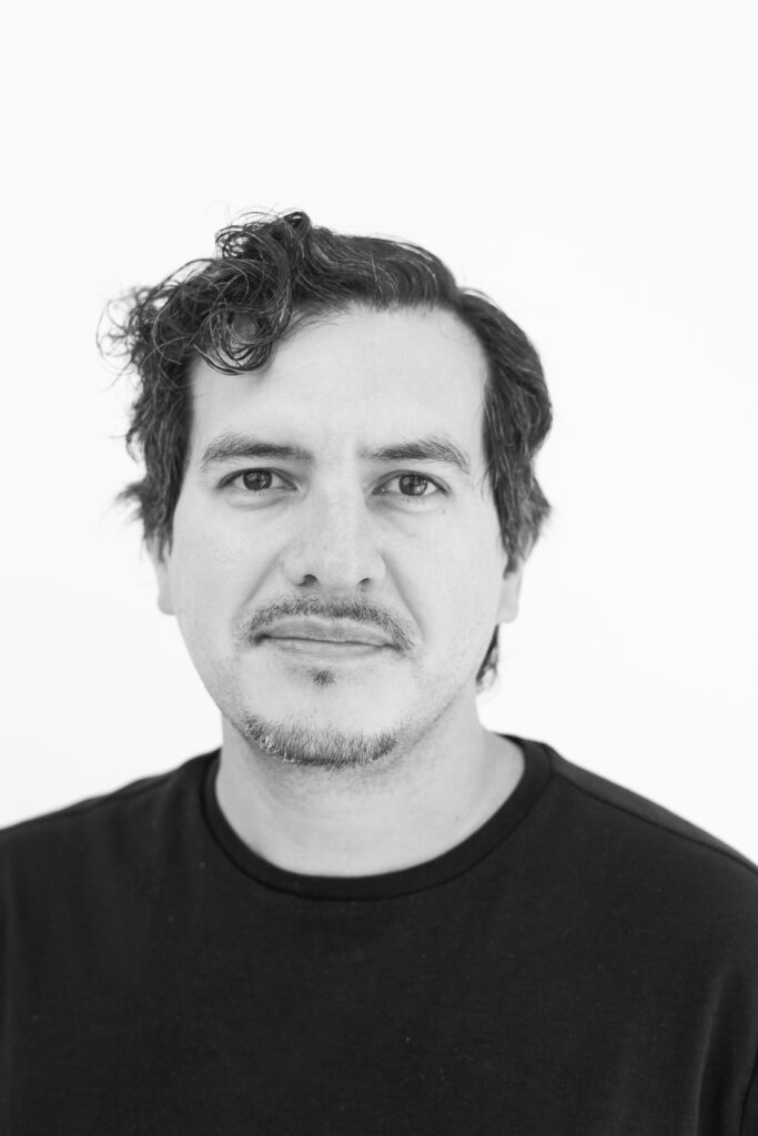 A black and white image of a person wearing a dark colored shirt with short hair that curls in the front, a mustache, and beard. They look towards the camera with a small smile.