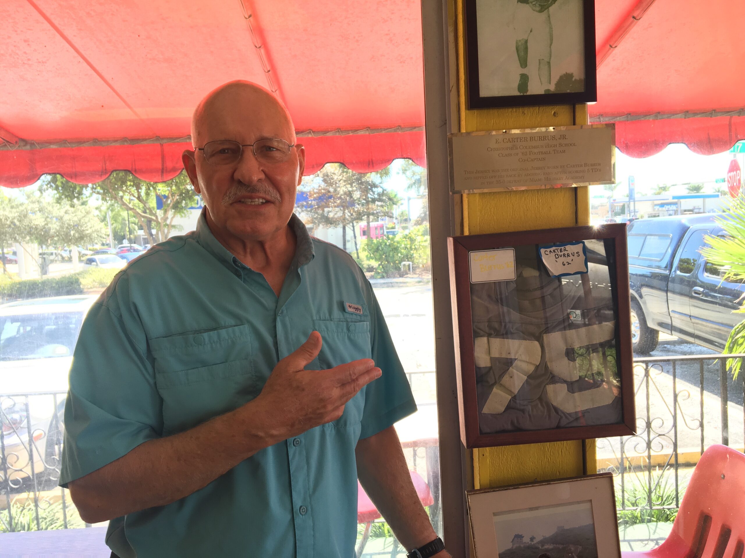 An older man, Carter Burrus, stands on an outdoor patio pointing at his framed jersey hung on a beam among other items. The framed jersey reads "75" and there s a sticker on the frame with his name.
