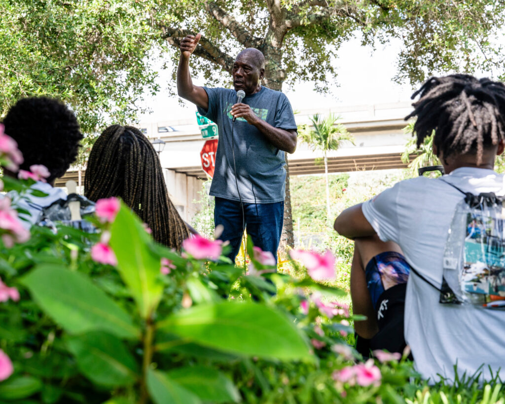 A person speaks into a microphone in a garden as three people look on.