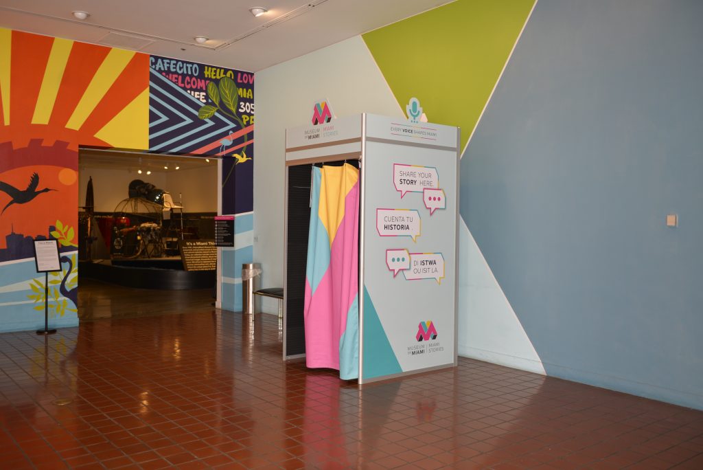 A standing booth with a colorful curtain in the front. the side panel of the booth reads "Every voice shapes Miami. Share your story here. Cuanta tu history. Di Istwa ou is it la."