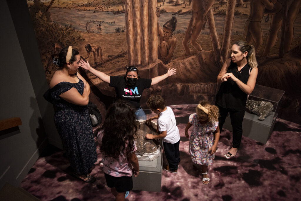 Group of people gather together on a tour of a museum exhibition. The tour guide has their hands outstretched while an ASL interpreter stands next to them.