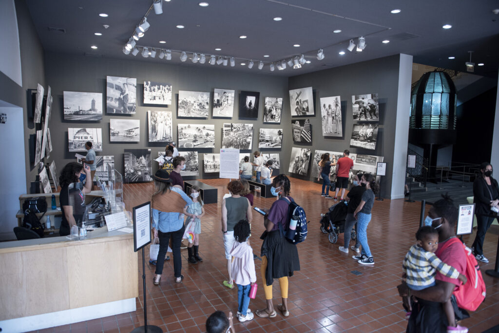 Group of people stand in a museum lobby. A staff member at the Visitor Services desk stands to greet them.