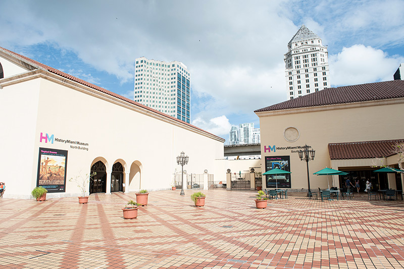 View of the Miami-Dade Cultural Plaza, with yellowy-beige and orange tiles, with Museum of Miami's two buildings in view. In the background is the Miami-Dade County Courthouse against a blue sky.
