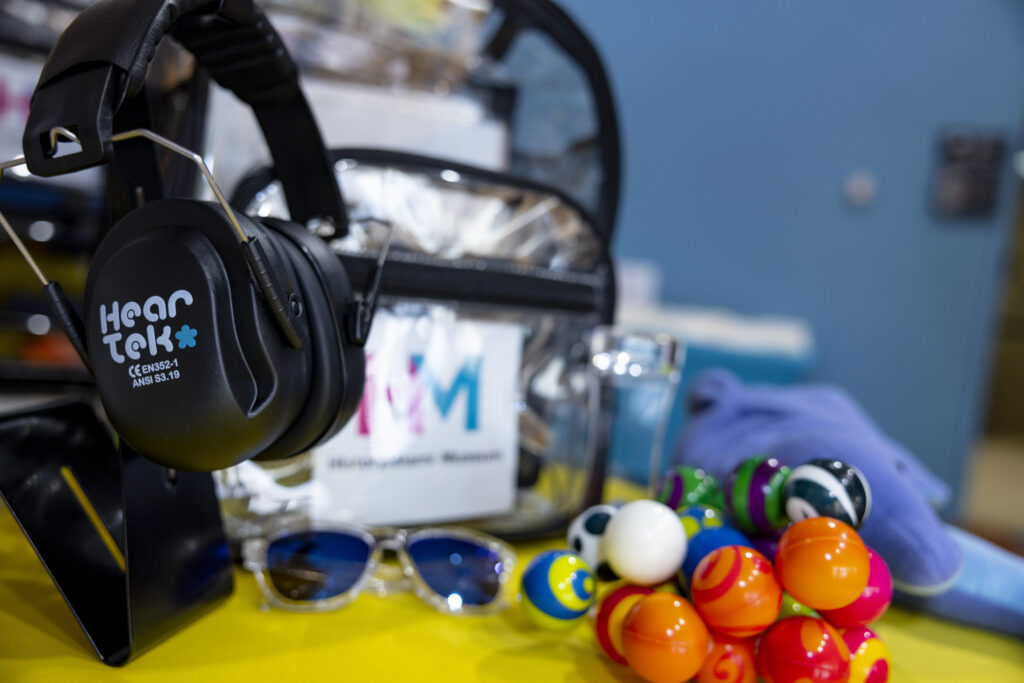 View of the museum's sensory backpack with support materials including noise reducing headphones, fidgets, sunglasses, and a weighted wrap in the shape of a dolphin.