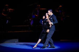 A man and woman are dancing tango. The woman stands with her back as he grabs her arm. They each have their left leg pointing forward.
