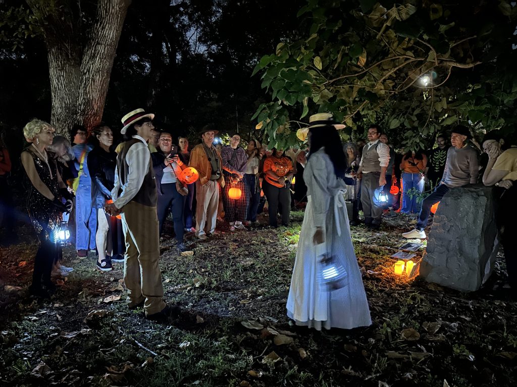 A group of tour participants stand in a cemetery at night. They look on to two performers in historical costume. Jack-o-lanterns and candles light up the scene.