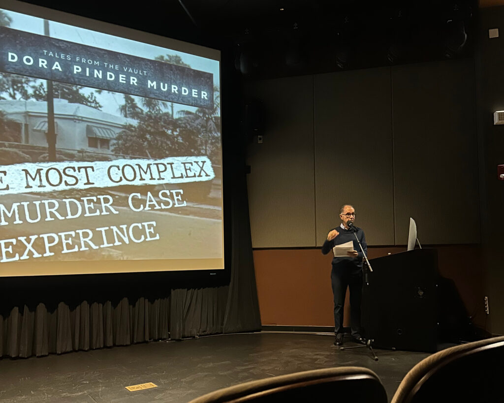 Dr. George stands on-stage in front of a projector screen that reads "most complex" and "murder case...experience."