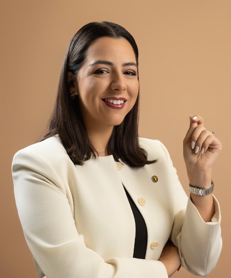 A person with shoulder length brown hair wearing a structured cream blazer, black blouse, silver watch, and small enamel pin smiles at the camera against a beige background.