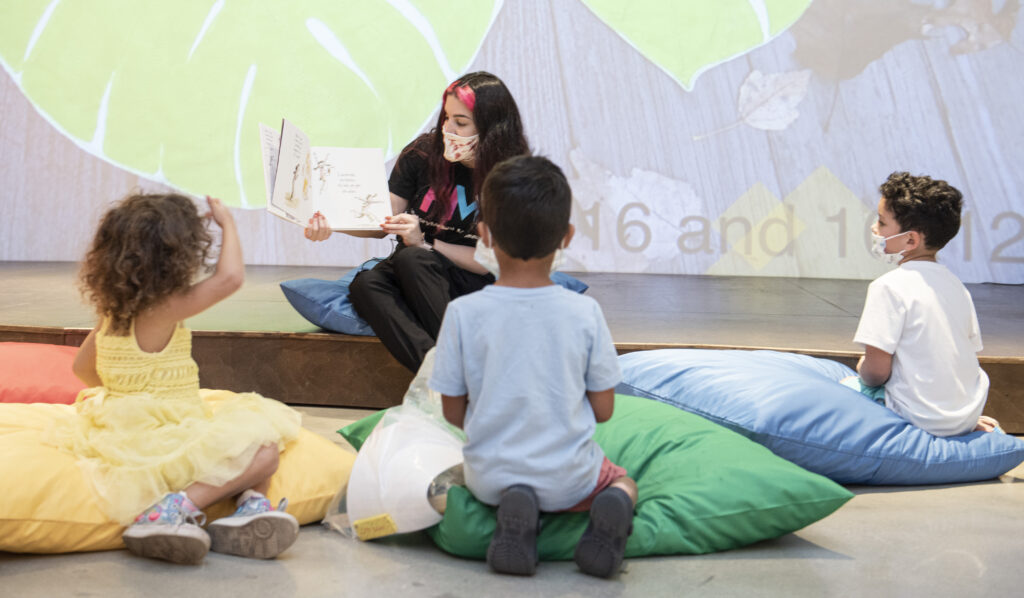 An adult seating on the floor in front of a room showing a book to three children seating on pillows