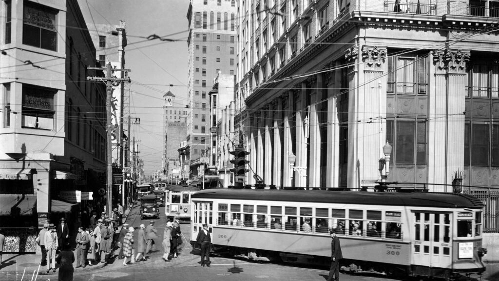 A black and white photograph of Downtown Miami in the 1930s. Trolley cars can be seen heading down a street alongside cars. A group of pedestrians are crossing the street with assistance from a police officer who stands in the middle of the road directing traffic.