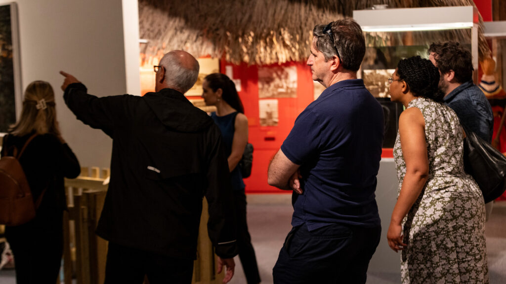A person points to an object inside of a Museum exhibition to a group of adults who look on.