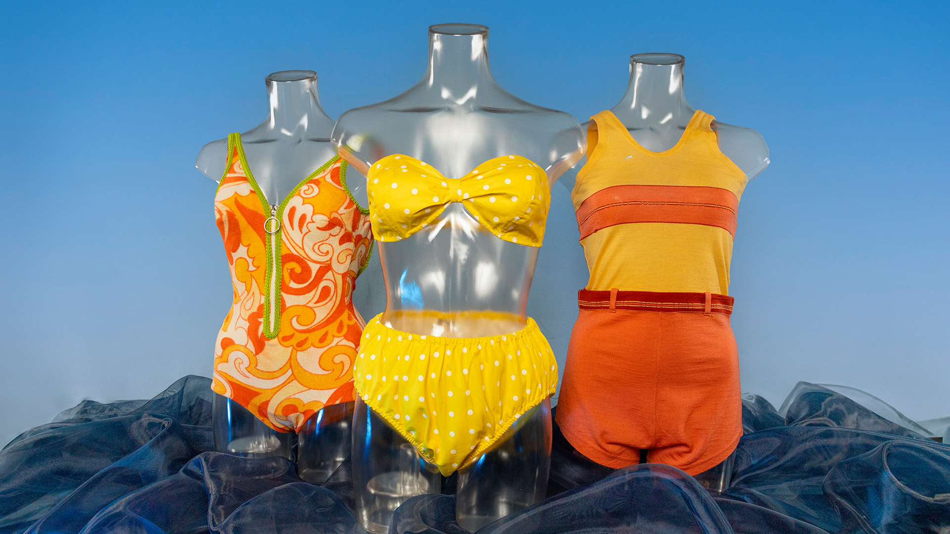 picture of three vintage swimsuits. The far left is orange with a green zipper, the middle is a yellow polka dot bikini, and on the right is an orange and yellow wool swimsuit.