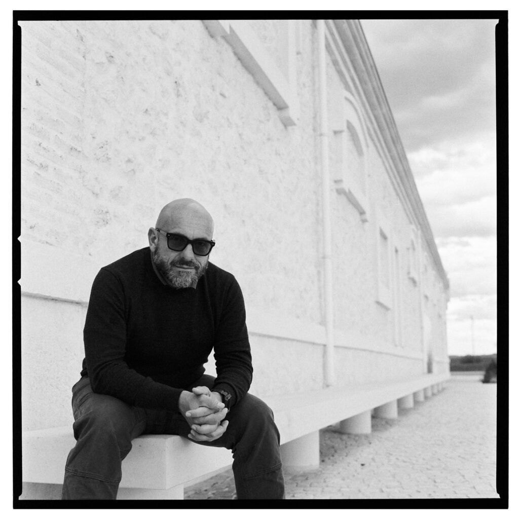 A person with a beard and dark sunglasses looks towards the camera as they sit with their arms on their legs and hands intertwined.