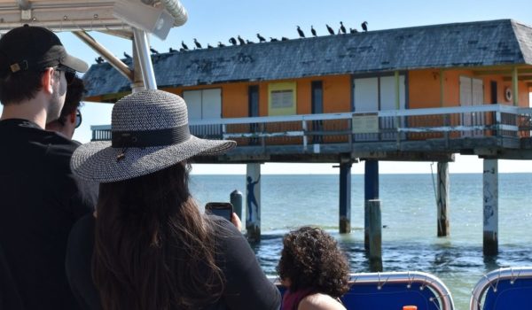 A group of people stand on a partially covered boat as they look towards an orange wooden house on stilts built out on the waters of Biscayne Bay.