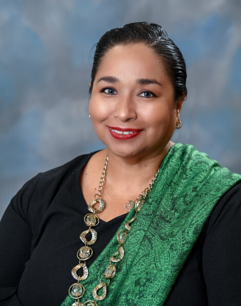 A person smiles at the camera. They wear their black hair pulled up with a black blouse and emerald green shawl draped across one shoulder.