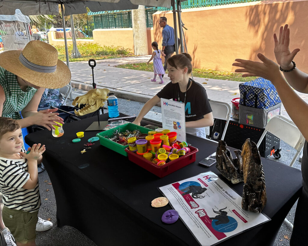A caregiver and child clap their hands as they stand in front of a table with play-doh and museum teaching collection items including a replica of a mastadon tooth and dire wolf skull.